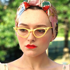 Op de afbeelding: Een vrouw met rode lippenstift draagt een roze en blauwe gebloemde hoofdband en gele cat-eye brillen.