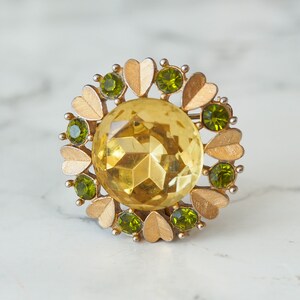 Peut inclure: Une broche dor&eacute;e avec une grande pierre jaune facet&eacute;e entour&eacute;e de petites pierres vertes et d'accents en forme de c&oelig;ur dor&eacute;s.
