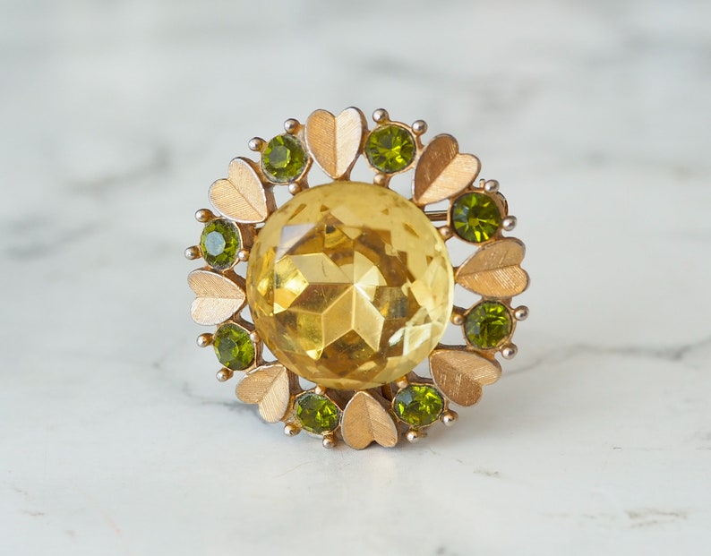 Peut inclure: Une broche dor&eacute;e avec une grande pierre jaune facet&eacute;e entour&eacute;e de petites pierres vertes et d'accents en forme de c&oelig;ur dor&eacute;s.