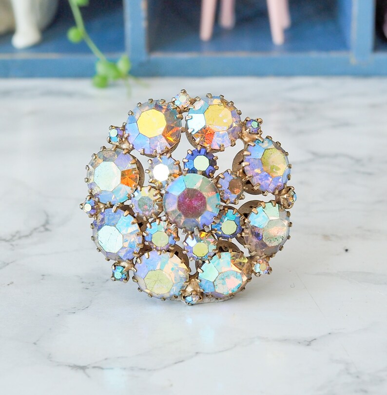 May include: A vintage brooch with a cluster of iridescent rhinestones in shades of blue, purple, and yellow. The rhinestones are set in a gold-toned metal.