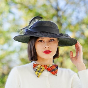May include: A woman wearing a black hat with a feather and a plaid bow tie. She is wearing a white sweater.