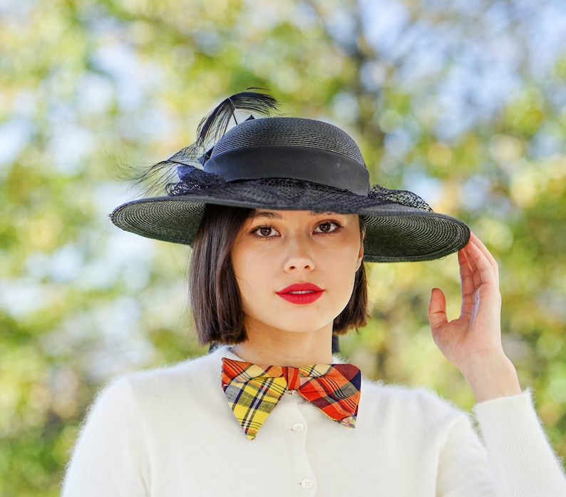 May include: A woman wearing a black hat with a feather and a plaid bow tie. She is wearing a white sweater.