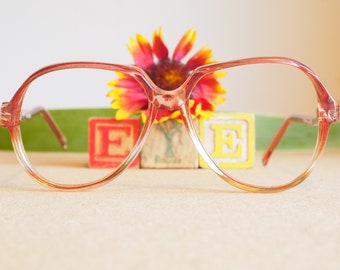 Könnte beinhalten: Rosa Plastik-Rahmenbrille mit klaren Gläsern. Die Brille liegt auf einem Tisch vor drei Holzklötzen, die "EYE" buchstabieren.