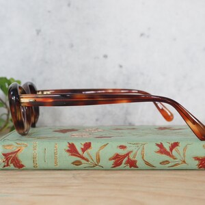 May include: Tortoiseshell glasses with gold accents resting on a vintage book with a green cover and gold leaf designs. The book title is "OUTRE-MER" and "LONGFELLOW".