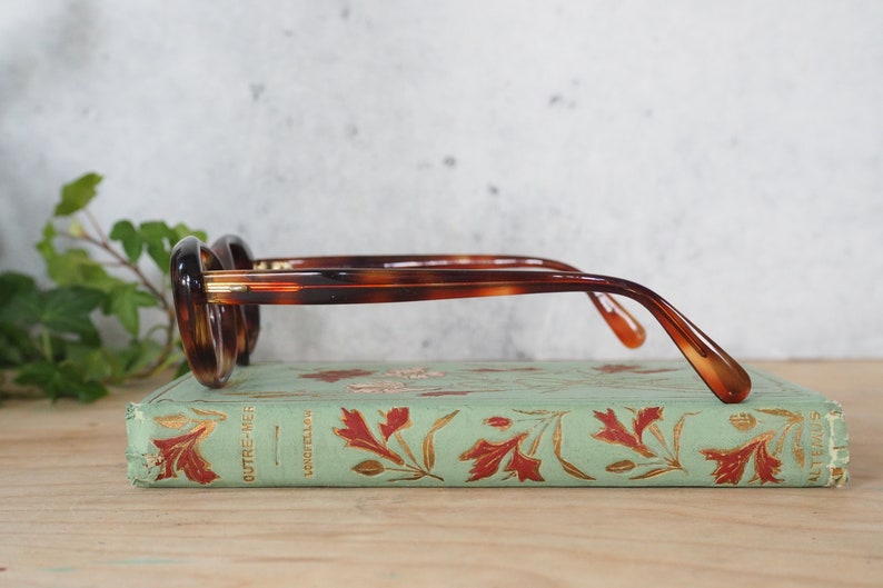 May include: Tortoiseshell glasses with gold accents resting on a vintage book with a green cover and gold leaf designs. The book title is "OUTRE-MER" and "LONGFELLOW".
