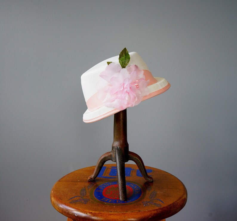 Op de afbeelding: Een witte hoed met een roze bloem en groene bladeren erop. De hoed staat op een houten standaard met een blauw-wit ontwerp.