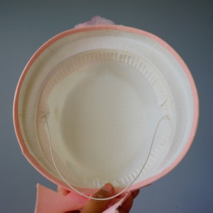 Op de afbeelding: Een roze hoed met een wit interieur en een draadframe. De hoed heeft een roze lint rond de basis.