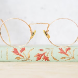 Peut inclure: Lunettes rondes en fil métallique dorées avec des verres transparents reposant sur un livre vert avec des motifs floraux rouges et des lettres dorées qui lisent "OUTRE-MER" et "LONDRES-LA"