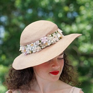 May include: A woman wearing a wide-brimmed straw hat with a band of white and pink flowers. The hat is beige and has a slightly rounded crown.