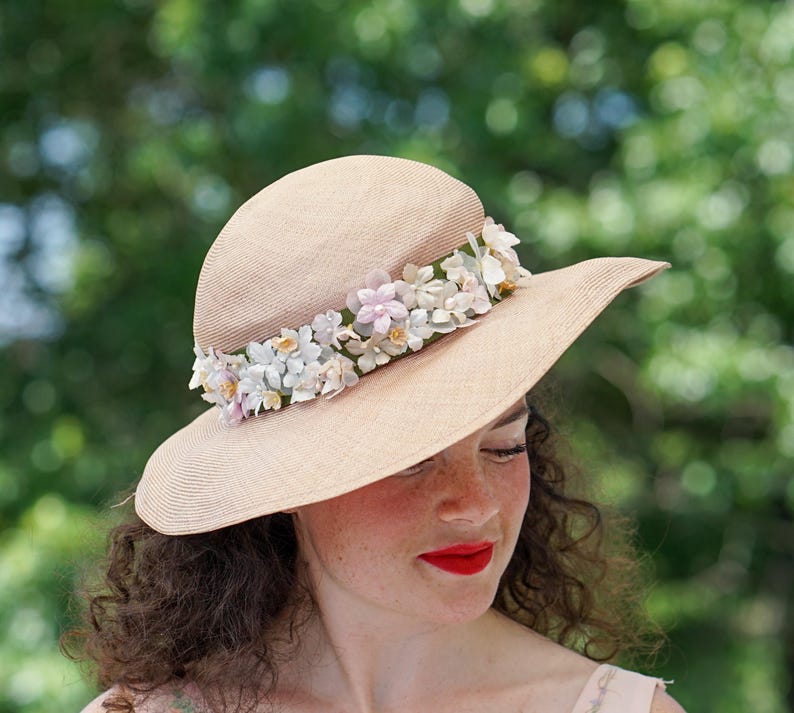 May include: A woman wearing a wide-brimmed straw hat with a band of white and pink flowers. The hat is beige and has a slightly rounded crown.