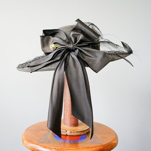 May include: A black straw hat with a large black satin bow and a jeweled pin. The hat is sitting on a wooden stand.