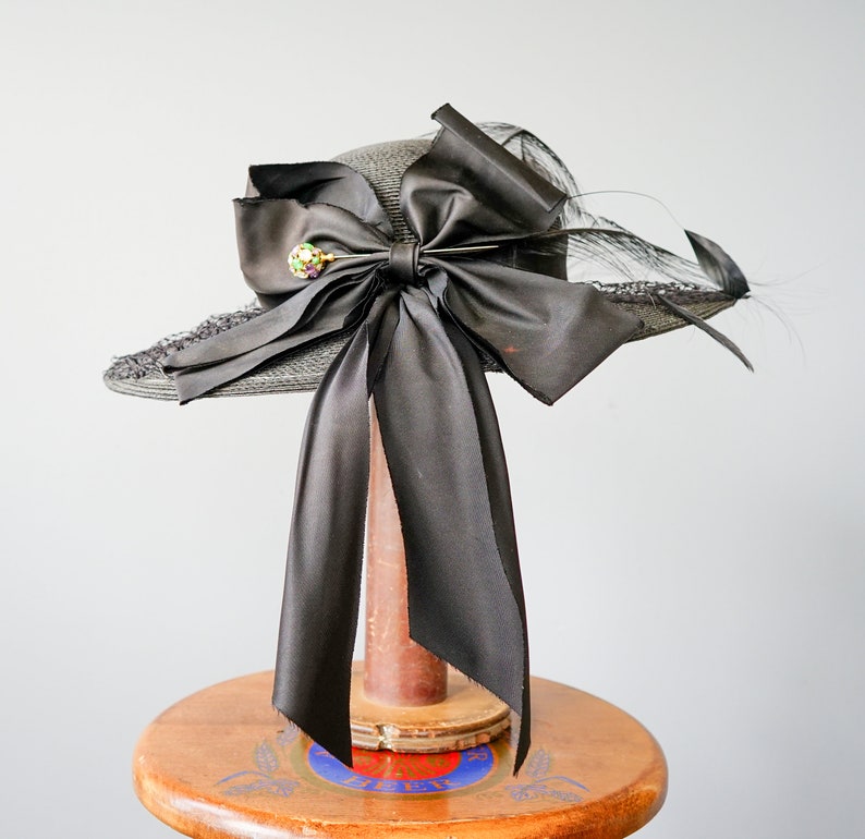May include: A black straw hat with a large black satin bow and a jeweled pin. The hat is sitting on a wooden stand.