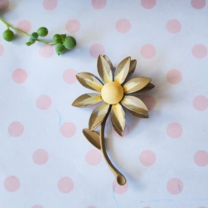 K&ouml;nnte beinhalten: Eine goldfarbene Brosche aus Metall in Form einer Blume mit gelbem Zentrum und Stiel. Die Brosche befindet sich auf einem wei&szlig;en Hintergrund mit rosa Punkten.