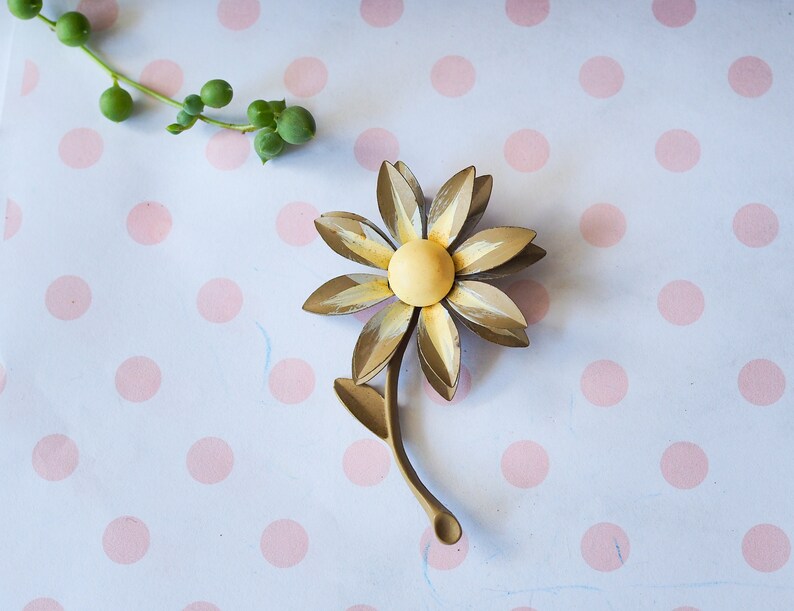 K&ouml;nnte beinhalten: Eine goldfarbene Brosche aus Metall in Form einer Blume mit gelbem Zentrum und Stiel. Die Brosche befindet sich auf einem wei&szlig;en Hintergrund mit rosa Punkten.