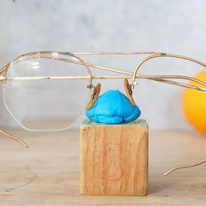 Peut inclure: Des lunettes à monture dorée avec des verres transparents reposent sur un objet bleu au sommet d'un bloc de bois avec le chiffre 5. Les lunettes ont un design classique à monture métallique. Un fruit orange est visible en arrière-plan.
