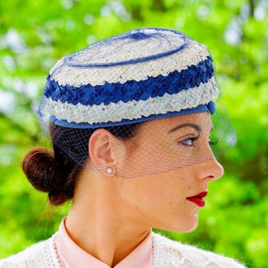 May include: A vintage hat with a blue and white striped design, with a matching veil. The hat is worn with a pink collared shirt and a white cardigan. The background is a blurred green.