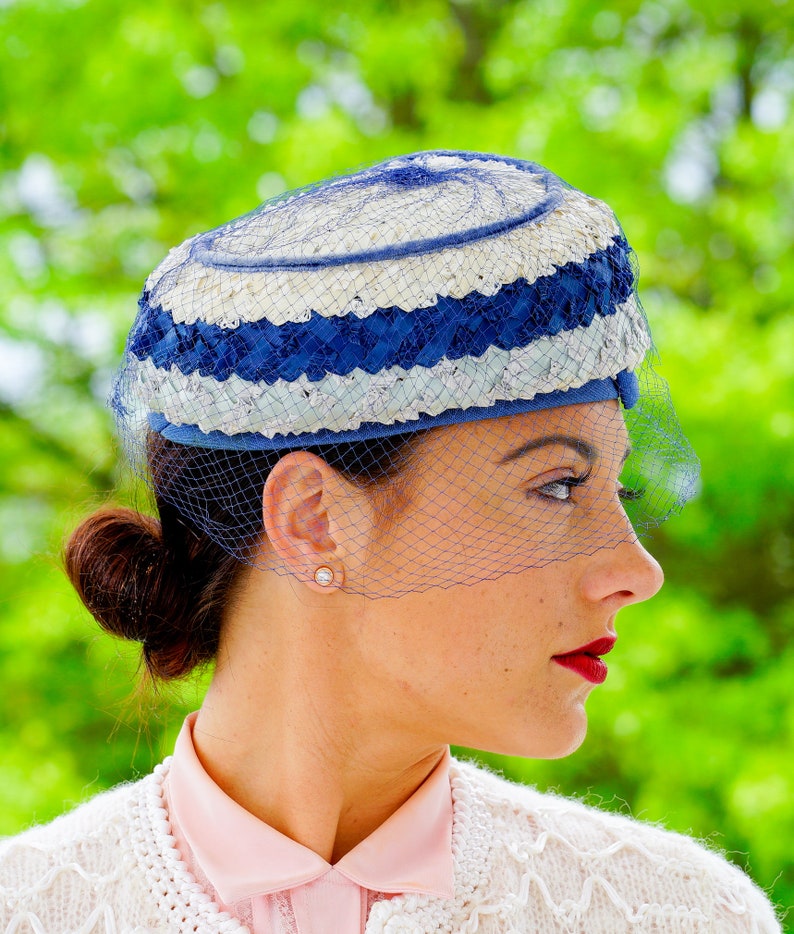 May include: A vintage hat with a blue and white striped design, with a matching veil. The hat is worn with a pink collared shirt and a white cardigan. The background is a blurred green.