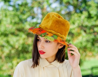 May include: A woman wearing a mustard yellow velvet hat with a band of colorful feathers. The hat has a wide brim and is tilted slightly to the side. The woman has short, dark hair and is wearing a white blouse.