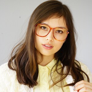 May include: A woman with long brown hair wearing a white cardigan and a yellow shirt. She is wearing large, round, tortoise-shell glasses.