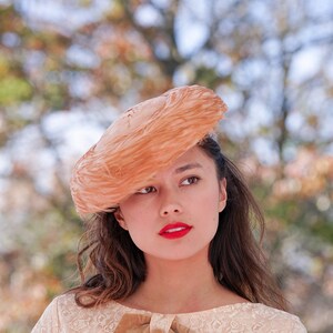 May include: A woman with long brown hair wears a peach-colored feathered hat. She is wearing a light beige lace dress with a brown ribbon.