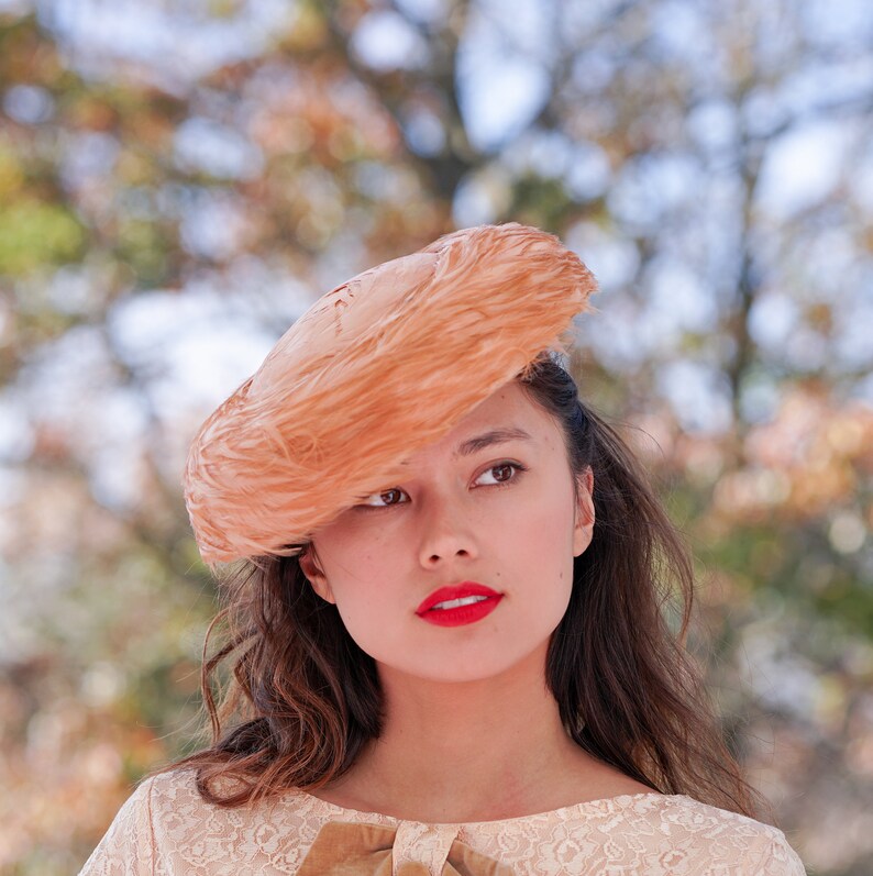 May include: A woman with long brown hair wears a peach-colored feathered hat. She is wearing a light beige lace dress with a brown ribbon.