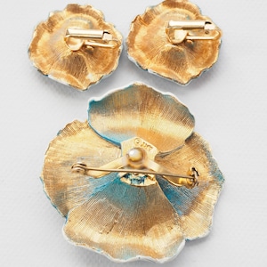 May include: A set of three gold-toned flower-shaped brooches with blue enamel accents. The brooch has a pin back and the earrings have clip-on backs. The brooch is marked "O.M.T."