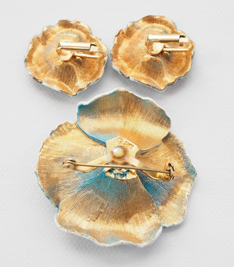 May include: A set of three gold-toned flower-shaped brooches with blue enamel accents. The brooch has a pin back and the earrings have clip-on backs. The brooch is marked "O.M.T."