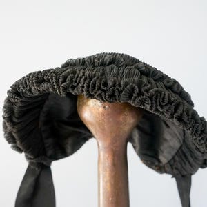 May include: A black, ruffled, fabric hat with a wide brim and two long ties. The hat is displayed on a wooden stand.