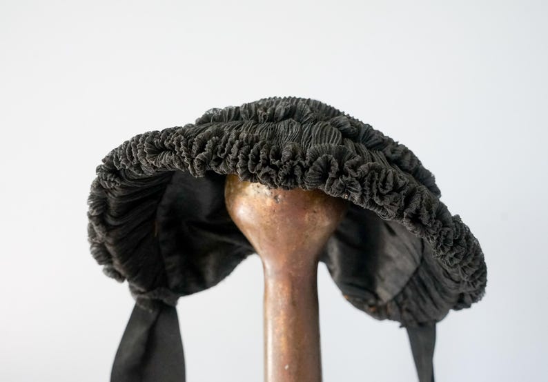 May include: A black, ruffled, fabric hat with a wide brim and two long ties. The hat is displayed on a wooden stand.