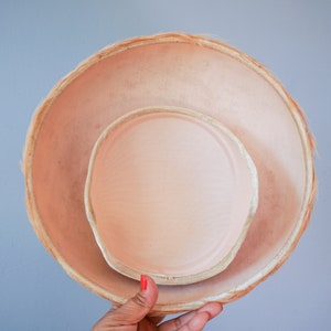 May include: A light pink hat with a wide brim and a smaller, inner hat. The hat is made of a woven material and has a soft, smooth texture.