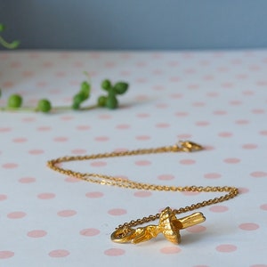 May include: A gold-colored necklace with a pendant shaped like a stylized flower or leaf. The necklace is displayed on a pink and white polka dot surface. The background includes greenery and a neutral-toned wall.