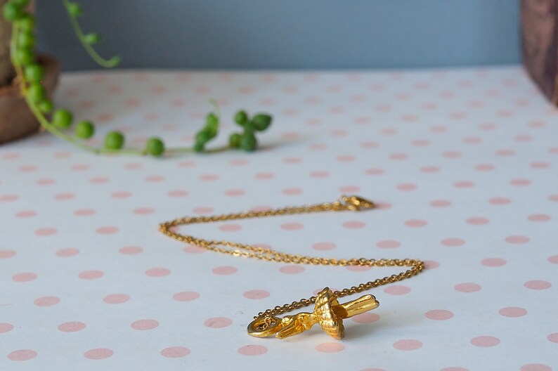 May include: A gold-colored necklace with a pendant shaped like a stylized flower or leaf. The necklace is displayed on a pink and white polka dot surface. The background includes greenery and a neutral-toned wall.