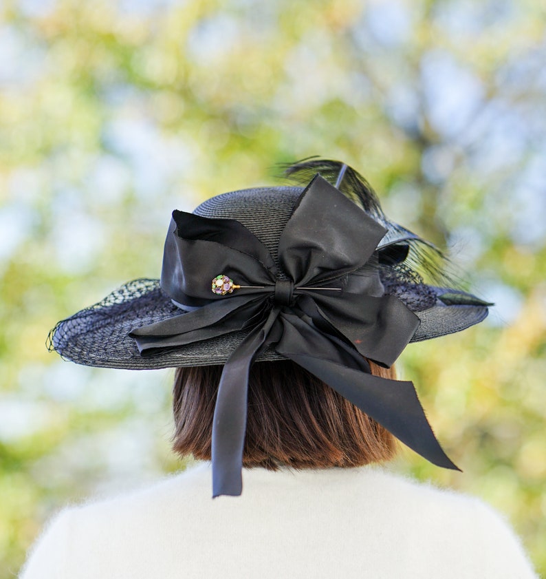 May include: A black hat with a large black bow and a small jeweled pin. The hat is made of straw and has a wide brim.