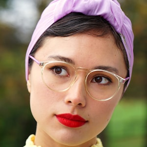 Può includere: Una donna con i capelli scuri che indossa un turbante color lilla chiaro e occhiali con montatura trasparente. Indossa un top di pizzo giallo e ha il rossetto rosso.