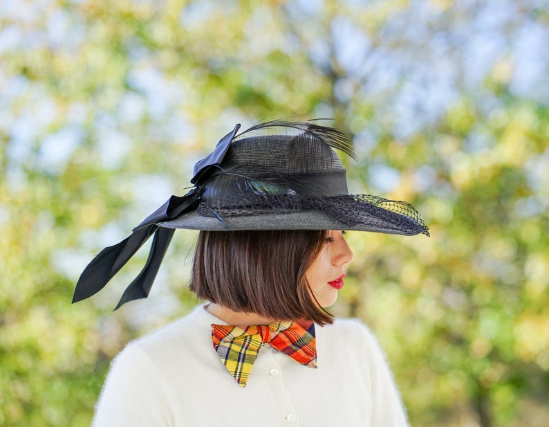 May include: A woman wearing a black hat with a black ribbon and feathers, a white sweater, and a plaid bow tie. The hat has a black mesh veil.