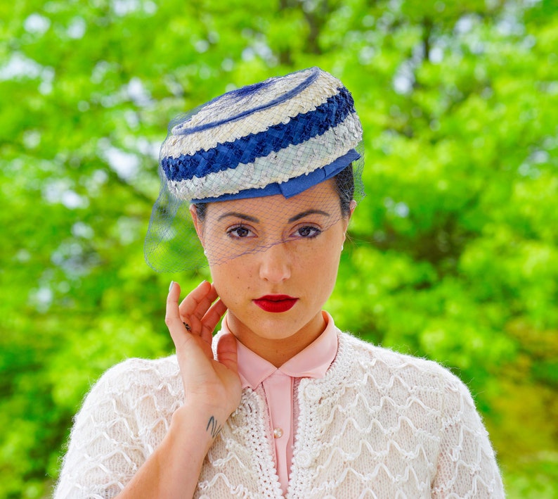 May include: A vintage hat with a blue, white, and cream striped design, with a fine blue veil. The hat is worn with a cream cardigan and a pink collared shirt. The background is a blurred green.