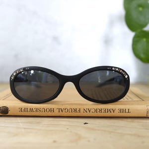 May include: Black oval sunglasses with decorative rhinestones along the top rim, resting on a vintage book titled "THE AMERICAN FRUGAL HOUSEWIFE". The lenses are dark, and the frame is matte black.