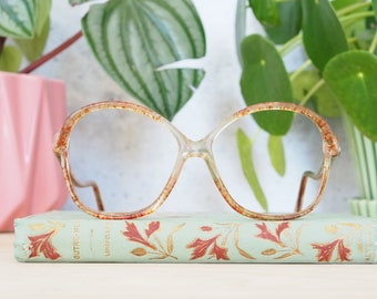 May include: A pair of vintage, tortoiseshell-colored eyeglasses with round lenses. The glasses are resting on a book with a floral design.