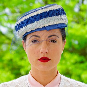 May include: A vintage hat in blue and white with a veil. The hat has a woven design with blue and white stripes and a blue ribbon. A sheer blue veil is draped over the face. The woman is wearing a pink collared shirt and a white cardigan.