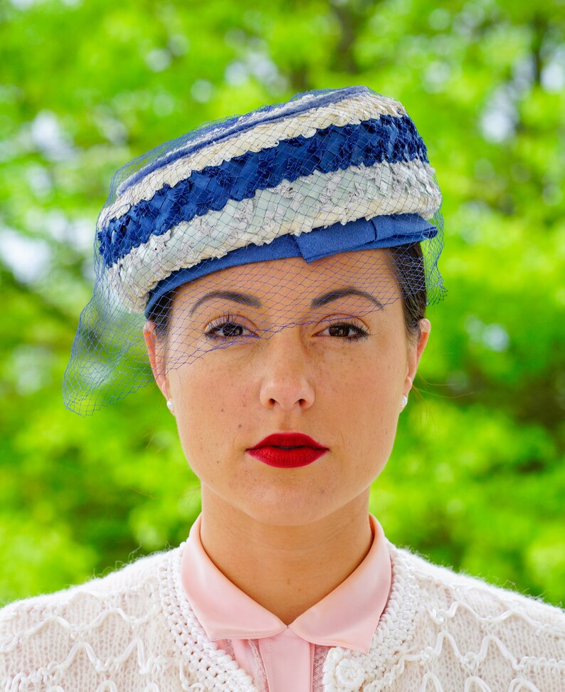 May include: A vintage hat in blue and white with a veil. The hat has a woven design with blue and white stripes and a blue ribbon. A sheer blue veil is draped over the face. The woman is wearing a pink collared shirt and a white cardigan.