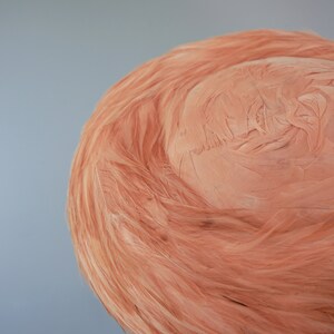 May include: A close-up of a light peach-colored, round, woven hat. The hat is made of a soft, fibrous material that resembles feathers.