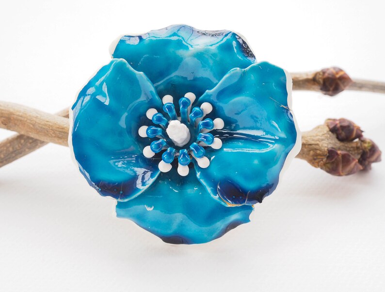 May include: A blue ceramic flower brooch with white and blue accents. The flower has a glossy finish and is shaped like a poppy.