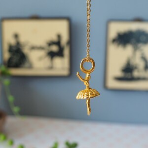 May include: A gold-colored ballerina pendant necklace. The pendant features a detailed ballerina figure with outstretched arms and a tutu. The necklace hangs from a delicate gold chain. Two framed silhouette art pieces are in the background.