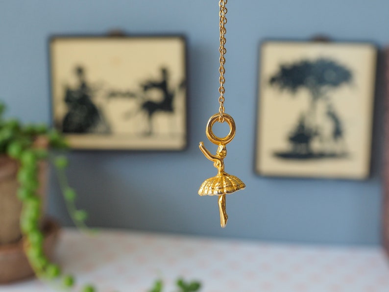 May include: A gold-colored ballerina pendant necklace. The pendant features a detailed ballerina figure with outstretched arms and a tutu. The necklace hangs from a delicate gold chain. Two framed silhouette art pieces are in the background.