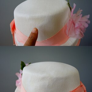 Op de afbeelding: Een witte strohoed met een roze lint en een roze bloemenversiering.