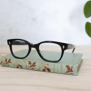 Op de afbeelding: Zwarte bril met een cat-eye vorm rust op een vintage boek met een vervaagde groene kaft en bloemenpatroon. De bril heeft een ruitvormig detail op de veren. De pagina's van het boek zijn zichtbaar.