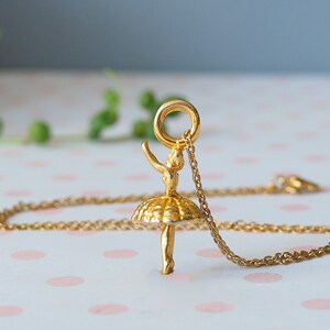 May include: A gold-colored ballerina pendant necklace. The pendant features a detailed ballerina figure with arms raised and a tutu. The necklace chain is also gold-colored. The background is a soft pink and white polka dot pattern.
