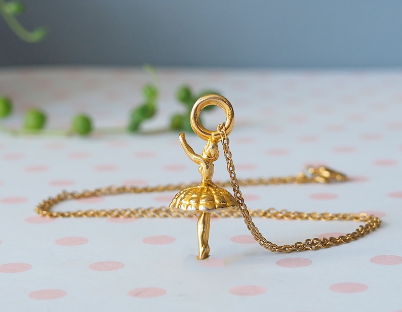 May include: A gold-colored ballerina pendant necklace. The pendant features a detailed ballerina figure with arms raised and a tutu. The necklace chain is also gold-colored. The background is a soft pink and white polka dot pattern.