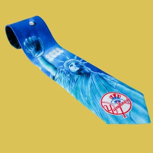 May include: A blue necktie featuring a graphic print of the Statue of Liberty holding a baseball glove, with the New York Yankees logo prominently displayed near the pointed tip. The design includes a baseball motif integrated into the team logo.