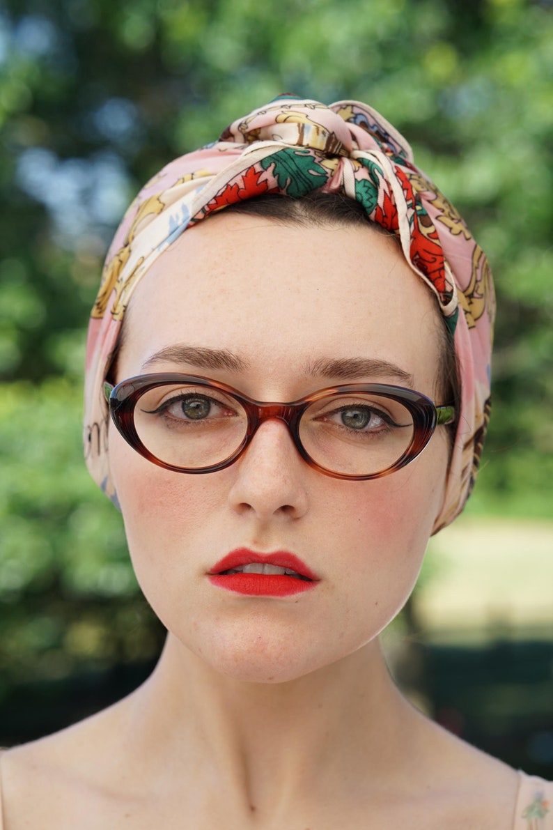 May include: A woman with red lipstick and brown cat-eye glasses is wearing a pink and green patterned headscarf.